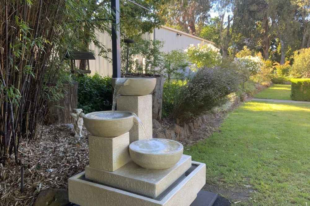 A three-tiered stone water fountain flowing in a lush green garden setting with sunlight-